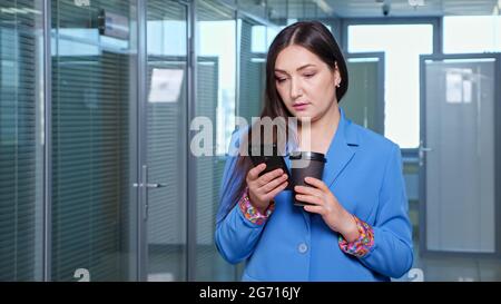 Brunette concentrata in giacca blu tipi su smartphone nero e tiene la tazza di caffè in piedi in sala tra gli uffici contro finestra, spazio di copia Foto Stock