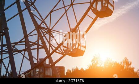 Le cabine vuote di un grande giro di divertimento in metallo in ferro ruota nel parco locale contro il cielo sopra le sagome degli alberi di sera dopo il tramonto Foto Stock