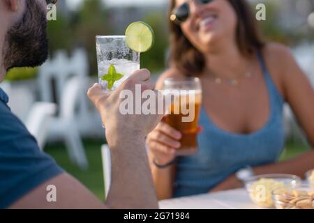 Una giovane coppia al caffè che beve bevande analcoliche lunghe ghiacciate divertendosi insieme. Mettere a fuoco il vetro con il mojito. Concetto di atmosfera estiva Foto Stock