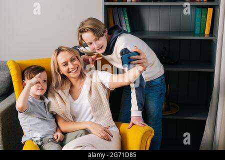 Ritratto di mamma allegra che ride prendendo selfie con due figli su smartphone seduti in poltrona gialla in un accogliente soggiorno. Foto Stock