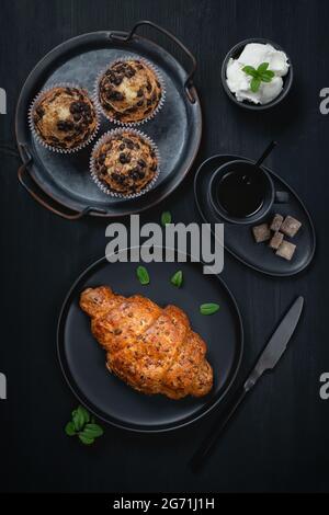 Tazza di caffè, muffin e croissant su tavolo nero in legno Foto Stock