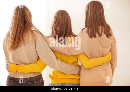 Vista posteriore di amici femminili anonimi in abiti casual che si abbracciano l'un l'altro su sfondo beige Foto Stock