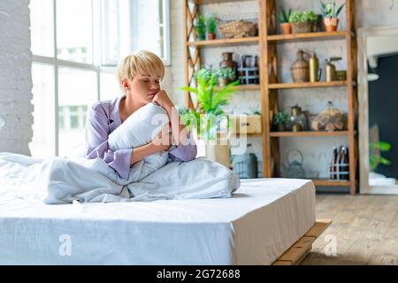 Donna bionda triste seduta sul letto la mattina e abbracciando il suo cuscino. Non ha avuto abbastanza sonno. Concetto di sonno sano. Foto Stock