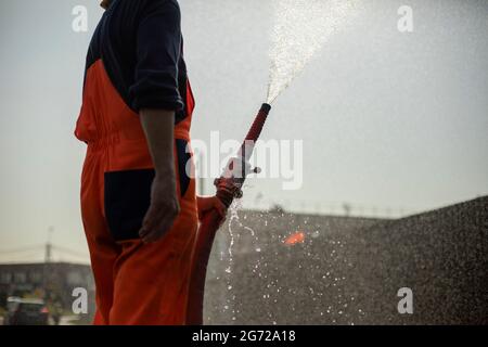 Un uomo versa l'acqua da un tubo flessibile. Una fontana di acqua da un idrante. Annaffiare la strada. Foto Stock