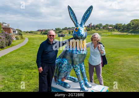 North Berwick, East Lothian, Scozia, Regno Unito, 10 luglio 2021. Il Big Hare Trail: Il sentiero inizia oggi e durerà 11 settimane con 10 gigantesche sculture di lepre dipinte a mano, ciascuna di un artista diverso, situate intorno alla città balneare. È organizzato da Leuchie House, una tregua di carità per le persone con disturbi neurologici. Nella foto: Hugh e Anne Barr sono venuti ad ammirare una delle lepri di Georgina Bown Foto Stock