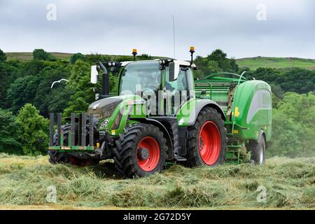 Produzione di fieno e insilato (agricoltore in cabina che guida un trattore agricolo verde sul campo, che tira l'imballatrice McHale, raccoglie l'erba secca tagliata) - Yorkshire Inghilterra UK Foto Stock