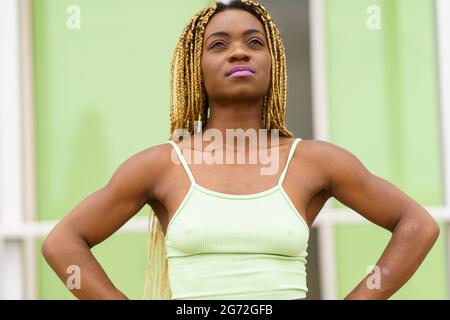 Donna con le mani in vita guardando orgogliosamente verso l'alto. Donna afroamericana con trecce gialle e abbigliamento sportivo verde chiaro che guarda con sicurezza verso l'alto. Foto Stock