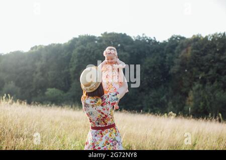 Madre e la sua piccola figlia che si divertono sul campo. Famiglia felice su un prato estivo al tramonto raggi. Madre tiene la sua figlia e la alza e gira Foto Stock