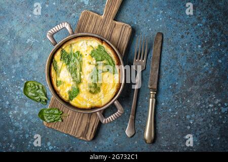 Vista dall'alto di gustose omelette con foglie di spinaci verdi in padella vintage con posate su tavola di legno e fondo di cemento blu Foto Stock