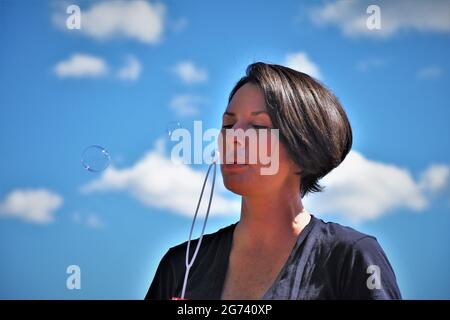 Donna caucasica dai capelli scuri che soffia bolle con cielo aperto dietro di lei, negli anni 30 Foto Stock