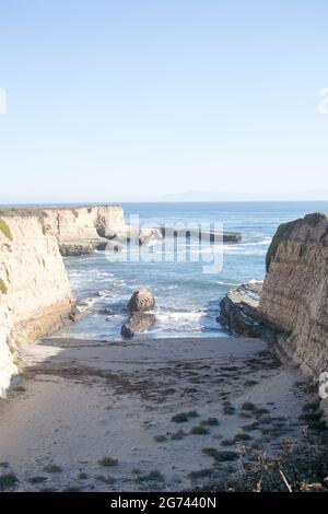 Piccola baia a scatola stretta circondata da alte scogliere striate che mostrano l'effetto dell'erosione. Spiaggia sabbiosa, panca di roccia offshore nell'oceano. Foto Stock