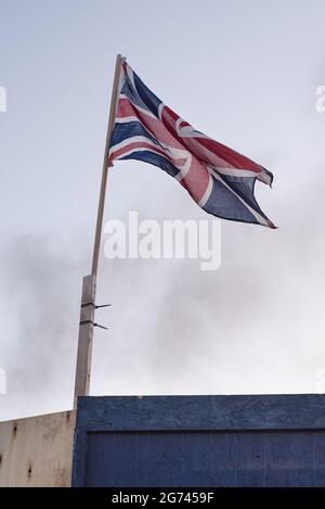 Belfast, Regno Unito. 10 luglio 2021. Una bandiera Jack Union apposta nell'area di Sandy Row e dietro di essa si vede del fumo. Per celebrare la battaglia del Boyne, i falò sono costruiti in vari quartieri protestanti in tutta Belfast, così come le strade e le case sono decorate con Union Jacks. I falò sono accesi la notte prima delle dodici sfilate di luglio, l'11 luglio. (Foto di Natalia Campos/SOPA Images/Sipa USA) Credit: Sipa USA/Alamy Live News Foto Stock