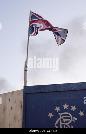 Belfast, Regno Unito. 10 luglio 2021. Una bandiera di Union Jack vista fissata alla parte superiore di un muro murale del Sandy Row Rangers Club nella zona di Sandy Row, con fumo visibile dietro di esso. Per celebrare la battaglia del Boyne, i falò sono costruiti in vari quartieri protestanti in tutta Belfast, così come le strade e le case sono decorate con Union Jacks. I falò sono accesi la notte prima delle dodici sfilate di luglio, l'11 luglio. (Foto di Natalia Campos/SOPA Images/Sipa USA) Credit: Sipa USA/Alamy Live News Foto Stock