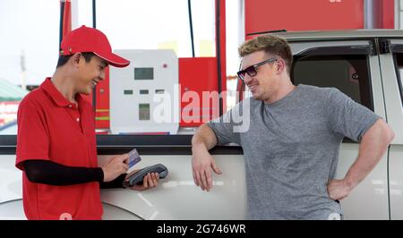 Distributore di benzina in rosso uniforme stand sorridente, strisciare mockup carta di credito tramite il terminale di pagamento. Autista caucasico con occhiali da sole in attesa accanto al bronzo Foto Stock