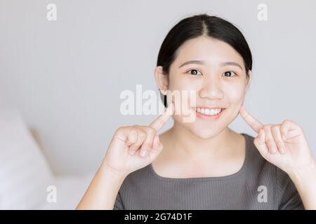 Bella giovane asiatica donna bianco bambino viso denti sani guardare il dito punto al viso per il concetto di cura della pelle del viso. Foto Stock