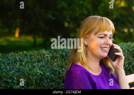 Defocalizzazione primo piano sorridente bionda caucasica che parla, parlando al telefono esterno, all'aperto. donna di 40 anni in blusa viola nel parco. pazienti adulti in pe Foto Stock
