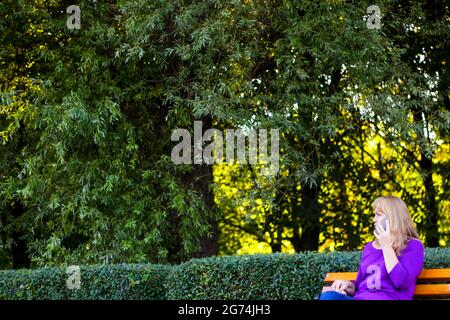 Defocus bionda caucasica donna che parla, parlando al telefono esterno, all'aperto. donna di 40 anni in blusa viola nel parco. Donne adulte che usano dispositivi mobili Foto Stock