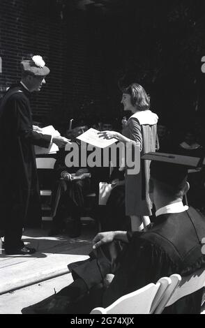 1964, storica, una cerimonia di laurea all'aperto, University of Southern California, USA. Un professore universitario su un palcoscenico in abbigliamento formale, un abito e testiera morbida, un cofano Tudor, con giovane donna, guardato da un uomo anche in abito e indossando sulla sua testa, un cappellino tradizionale laureato, un cappello a forma quadrata appiattito noto come un mortarboard. Foto Stock