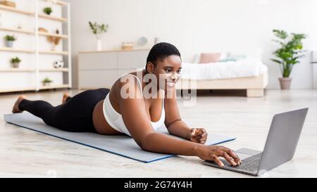 Donna nera sovrappeso sdraiata su un tappetino sportivo, utilizzando un computer portatile, comunicando con un allenatore personale o guardando un video di fitness Foto Stock