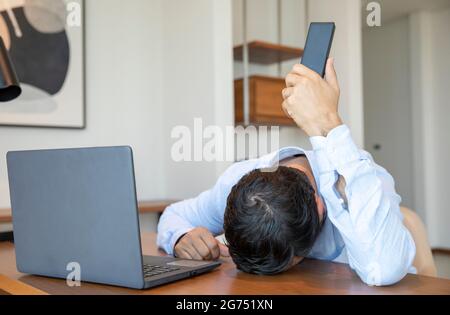 Frustrato uomo di mezza età che lavora da casa, di fronte al laptop, tenendo un telefono lontano dalle orecchie Foto Stock
