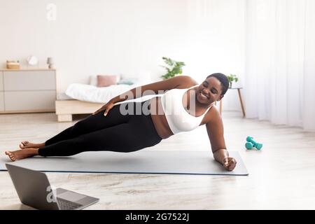 Sport in linea per perdita di peso. Donna nera in sovrappeso che fa esercizi davanti al computer portatile, in piedi nella tavola laterale a casa Foto Stock