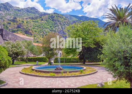 Montenegro, città di Kotor. Piazza con fontana vicino alla Città Vecchia di Cattaro e mura di antiche fortificazioni. Montagne e strada per fortezza in lontananza Foto Stock