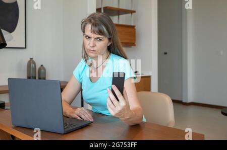 donna di mezza età che lavora da casa, infastidita, mantenendo il suo telefono lontano Foto Stock