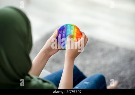 Vista ritagliata della ragazza teenage musulmana in hijab giocare con antistress POP IT giocattolo a casa, closeup delle mani Foto Stock