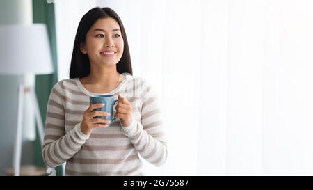 Sorridente donna asiatica bere caffè mentre in piedi vicino finestra a casa Foto Stock