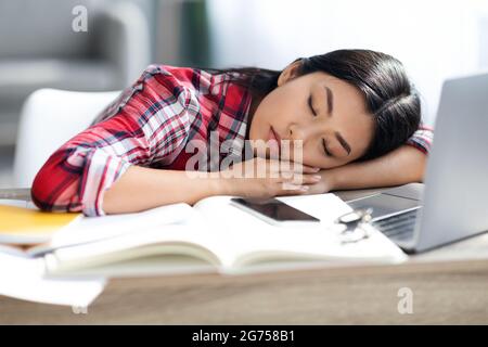 Esausta studentessa asiatica dormendo alla scrivania con libri e laptop Foto Stock