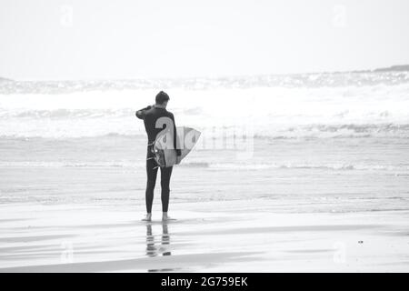 I surfisti di tutte le età portano tavole da surf sul bordo dell'acqua della spiaggia di Llangennith sulla penisola di Gower. Immagine monocromatica che raffigura attività all'aperto Foto Stock