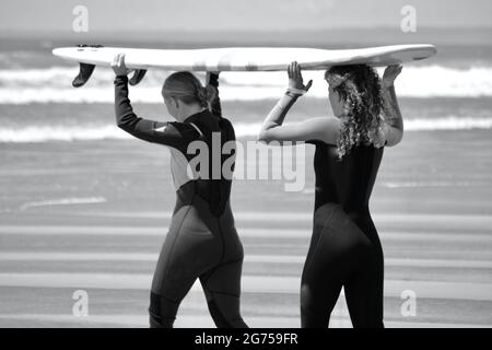 I surfisti di tutte le età portano tavole da surf sul bordo dell'acqua della spiaggia di Llangennith sulla penisola di Gower. Immagine monocromatica che raffigura attività all'aperto Foto Stock