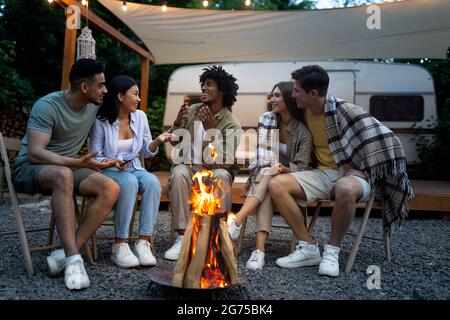 Felici amici multinazionali seduti vicino al falò, che si comunicano tra loro, godendo di una calda serata estiva accanto al camper Foto Stock