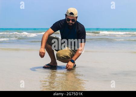 Il giovane al mare raccoglie conchiglie sulla spiaggia, tramonto, vacanze estive in mare o in oceano. Un ragazzo sulla spiaggia che raccoglie conchiglie. Foto Stock