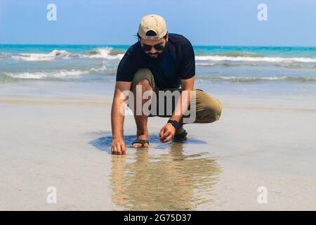 Il giovane al mare raccoglie conchiglie sulla spiaggia, tramonto, vacanze estive in mare o in oceano. Un ragazzo sulla spiaggia che raccoglie conchiglie. Foto Stock