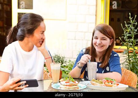 Felice giovane donna multirazziale amici seduti a tavola con panini e bevande fredde sulla terrazza del caffè durante l'orario di pranzo Foto Stock