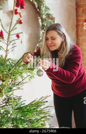 Vista laterale della donna adulta in rosso maglione appeso decorazioni su rami di conifere albero il giorno di Natale a casa Foto Stock