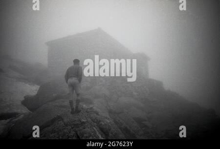 1935, storico, un escursionista maschile fuori nei Sudetes o Sudeten montagne in piedi su rocce, guardando attraverso la nebbia al profilo di un rifugio in pietra o rifugio di montagna, Cecosolvakia. Tali edifici offrono rifugio e rifugio a escursionisti ed escursionisti, quando si camminano sulla catena montuosa dei Sudeti, che sono la parte più alta del massiccio, noto come il Massiccio Boemo e che l'area, un tempo conosciuta come Sudetenland - annessa dalla Cecoslovacchia dalla Germania nel 1938 - è stata chiamata dopo. Foto Stock