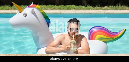 Vista panoramica di un giovane uomo che indossa occhiali da sole bevendo limonata in un anello gonfiabile unicorno in una piscina. Concetto estivo. Foto Stock