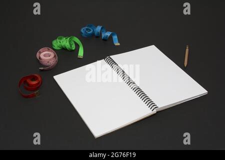 Un'immagine closeup di un notebook bianco con una matita e centimetri multicolore su uno sfondo nero Foto Stock