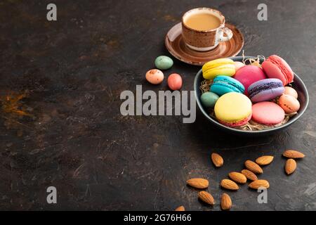 Macaroni multicolore e uova di cioccolato in ciotola di ceramica, tazza di caffè su fondo nero di cemento. Vista laterale, spazio copia, vita still. Prima colazione, m Foto Stock