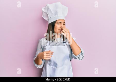 Giovane bella donna che indossa uniforme da cuoco professionale e cappello che odora qualcosa di stinoso e disgustoso, odore intollerabile, trattenendo il respiro con finge Foto Stock