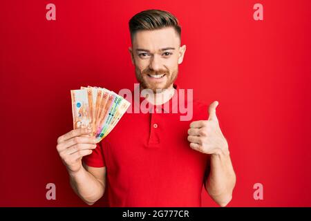 Giovane uomo rosso che tiene le banconote di peso filippino sorridente felice e positivo, pollice su facendo eccellente e segno di approvazione Foto Stock