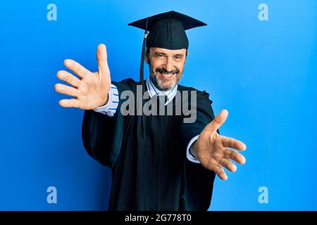 Uomo ispanico di mezza età con cappuccio di graduazione e abito da cerimonia guardando la macchina fotografica sorridente con le braccia aperte per abbraccio. Espressione allegra abbracciando h Foto Stock