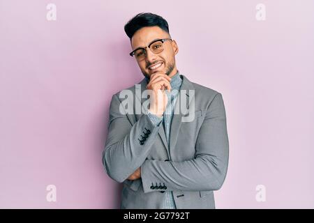 Giovane arabo che indossa abiti da lavoro sorridente guardando fiducioso la macchina fotografica con le braccia incrociate e la mano sul mento. Pensare positivo. Foto Stock
