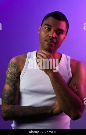 Ritratto verticale di un uomo di razza mista tatuato che guarda la fotocamera mentre si posa su sfondo viola in studio, copia spazio Foto Stock