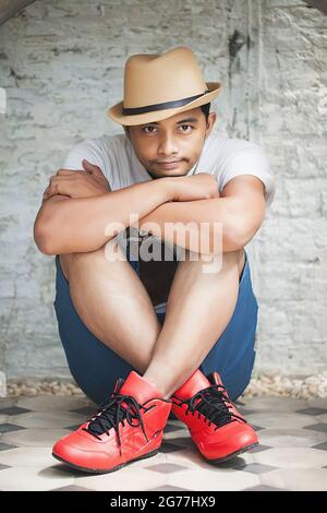 Attraente uomo hugging himself.in uno stile seduta position.Dress con colori impressionanti, scarpe rosse, pantaloncini blu. Foto Stock