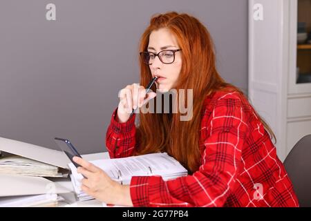 Donna con capelli rossi seduta alla scrivania con file ad anello che guardano il telefono cellulare in mano Foto Stock