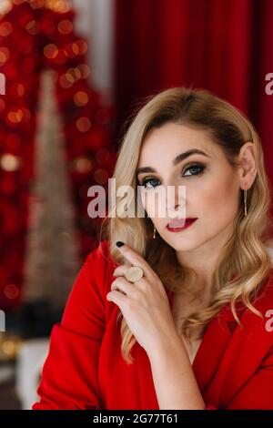 Ritratto giovane bella donna bionda in rosso sullo sfondo di luci bokeh di un albero di Natale. Felice anno nuovo 2021. Messa a fuoco selettiva e sfocata Foto Stock