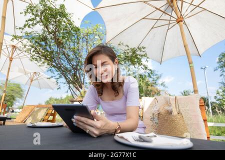 La bella donna di mezza età seduta in un negozio di alimentari e lavora con un computer tablet mentre attende il cibo con un bel cielo blu sullo sfondo Foto Stock